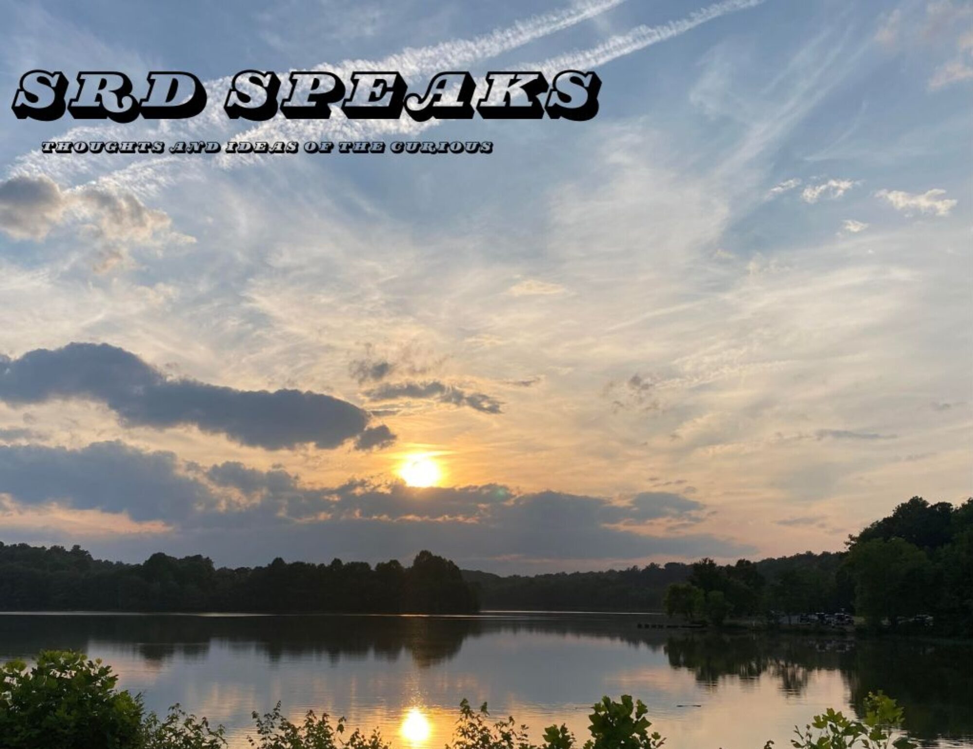 A sunset over a wooded lake area. The sunset is reflecting onto the lake as well as the clouds surrounding the sun. There is also some shrubbery and green trees surrounding the lake. In the top righthand corner, there are outlined words with the larger text saying, "SRD SPEAKS" and the smaller text saying, "thoughts and Ideas of the curious".